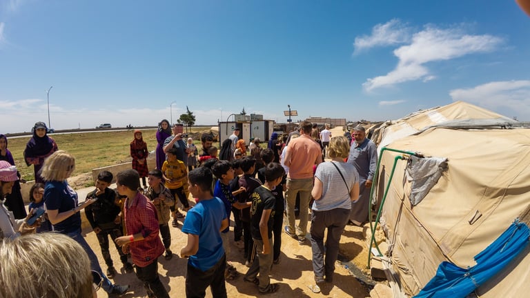 A group of people, including children and adults, gather near tents in an open field under a clear sky, receiving aid from visiting helpers
